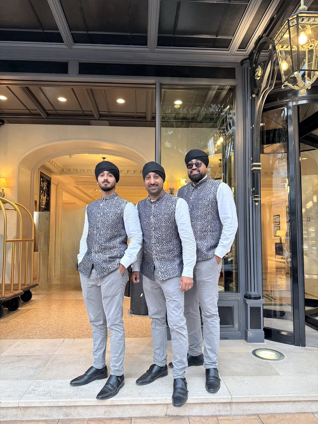 Three dhol players in patterned waistcoats and light trousers outside a hotel