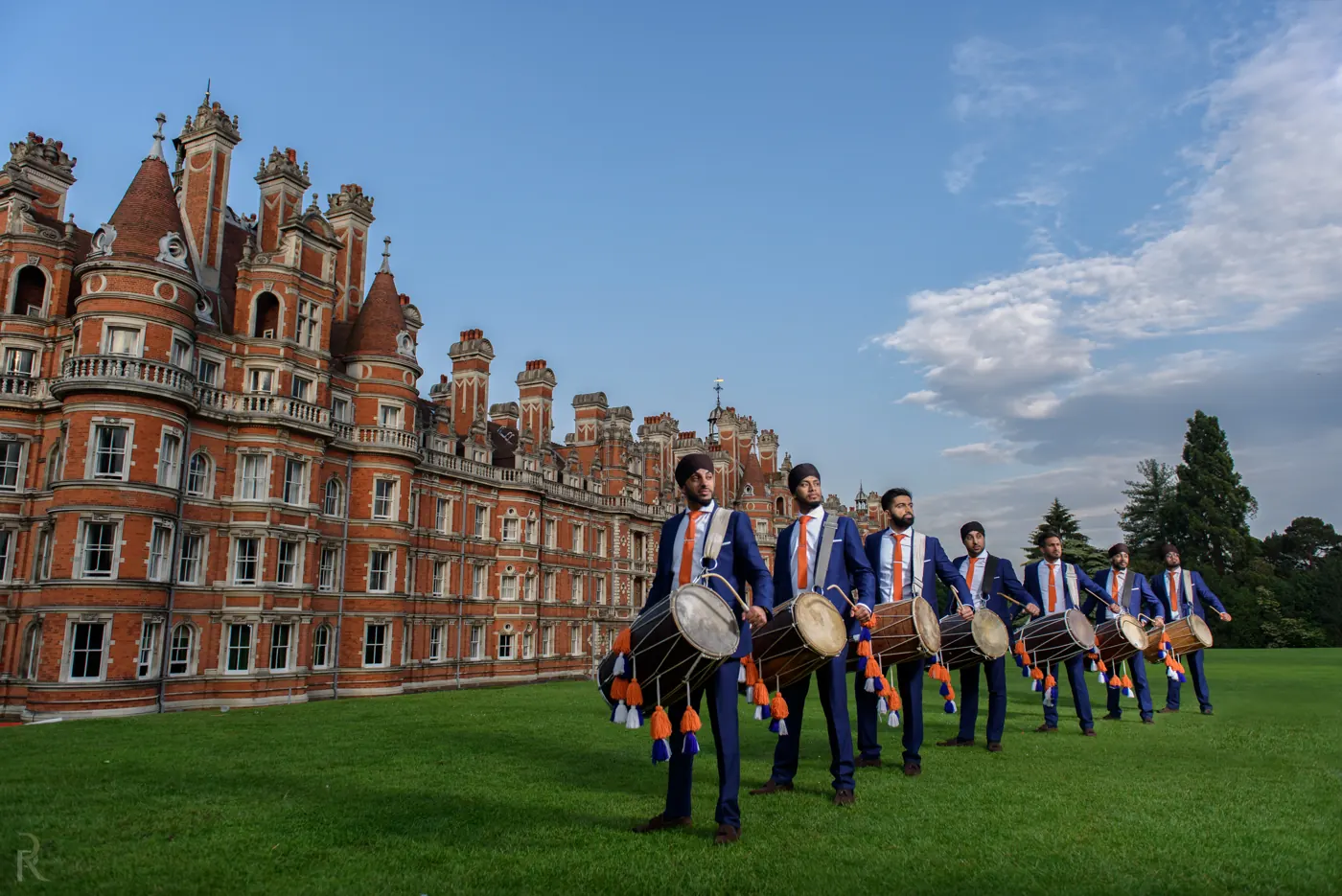 Dhol players in navy suits carrying dhols on the lawn in front of Royal Holloway, Egham