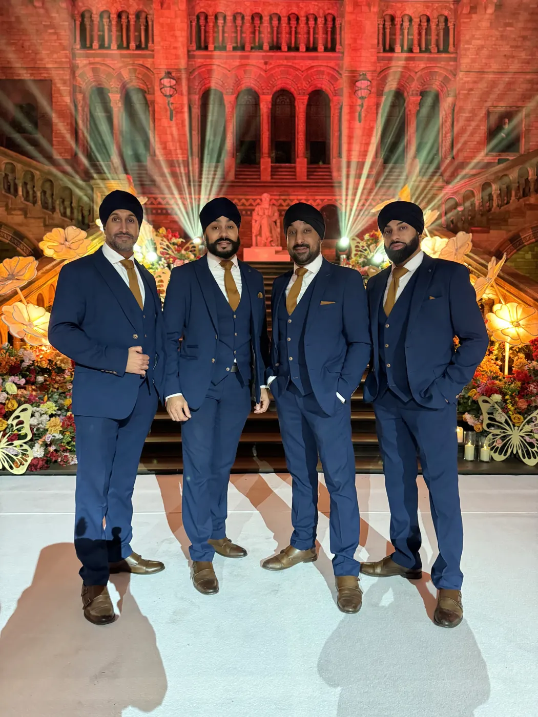 Four dhol players in navy three-piece suits in front of the lit Natural History Museum