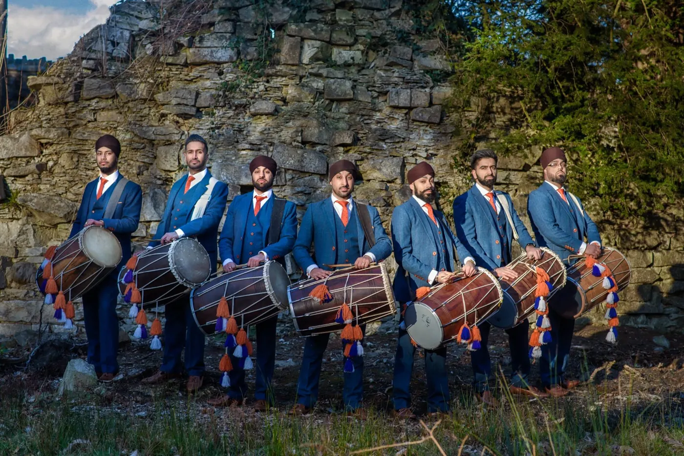 Seven dhol players in navy three-piece suits holding dhols in front of a rock wall at Virginia Water