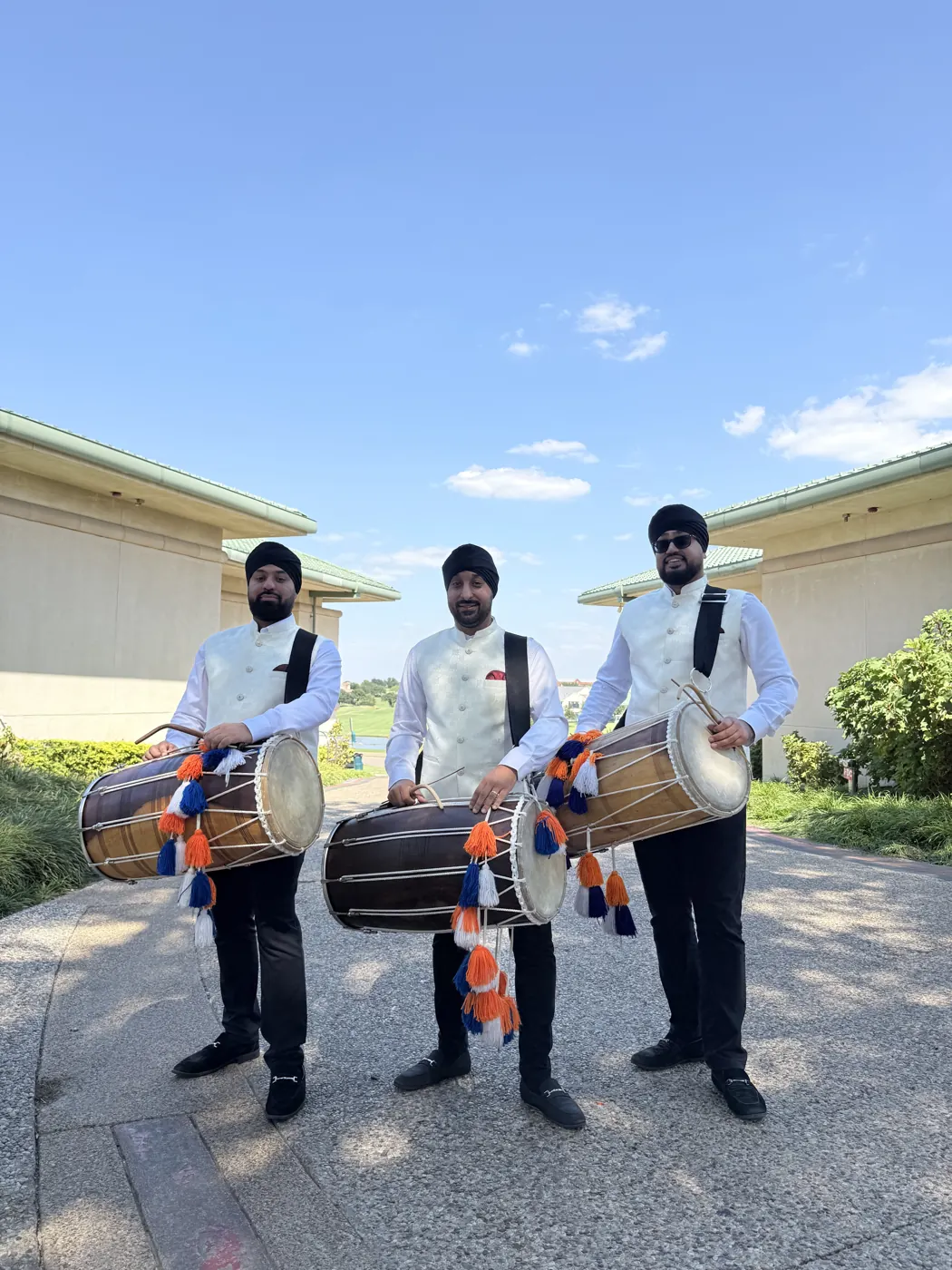 Three dhol players in white sherwani waistcoats and black trousers holding dhols outdoors